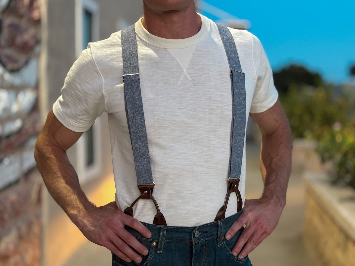 A man wearing a white crewneck t-shirt and blue denim suspenders with brown leather accents, standing outdoors with his hands on his hips.