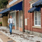 A man walks through a historic downtown wearing Espresso Linen Suspenders with black leather button-on attachments, paired with a vintage white Henley and classic denim. Handmade in Texas by Stratton Suspender Co., perfect for weddings, formalwear, and heritage-inspired everyday menswear.