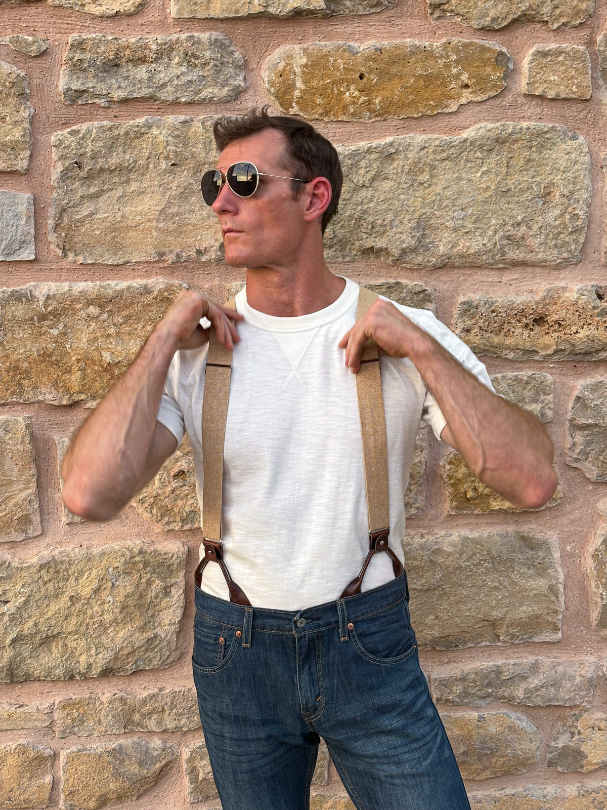 A man wearing round sunglasses, a white t-shirt, tan suspenders, and blue jeans, posing in front of a rustic stone wall.
