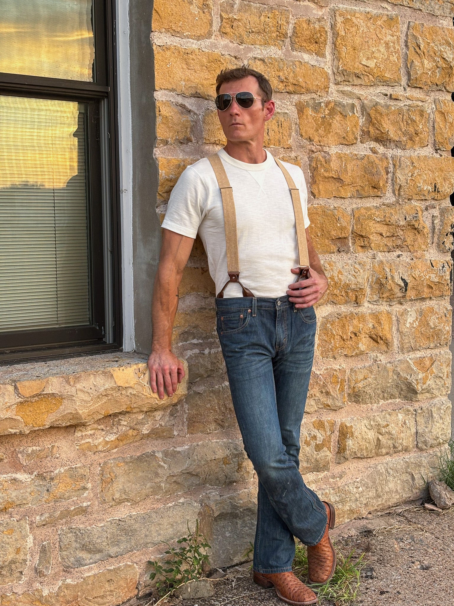 A man wearing beige suspenders, a white t-shirt, blue jeans, and brown boots leans against a rustic stone wall next to a window.