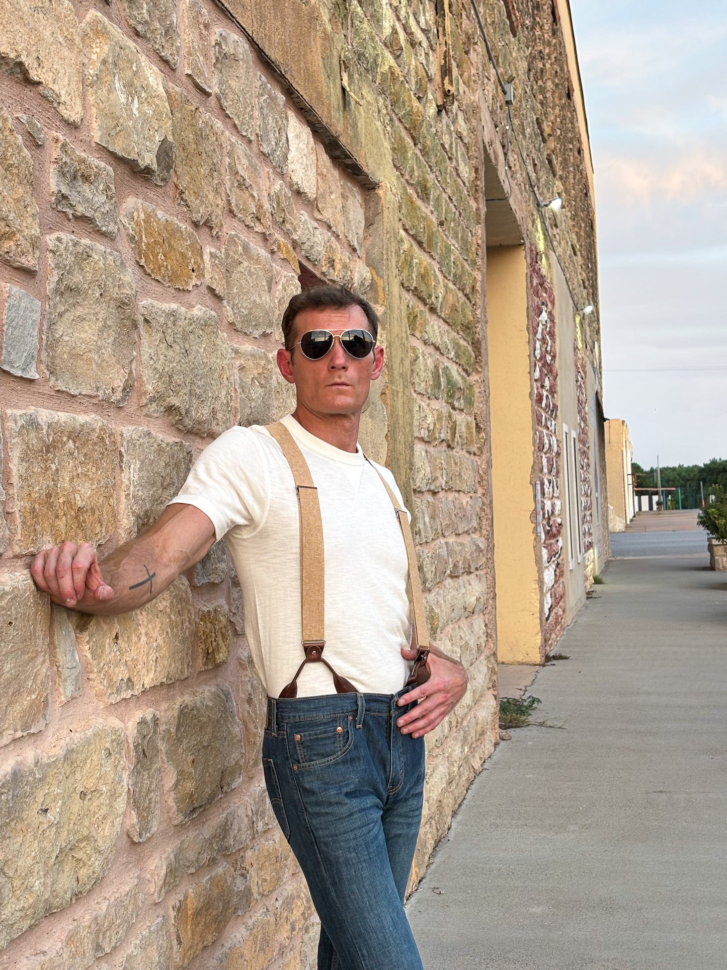 A man wearing a cream ribbed t-shirt, brown leather suspenders, and blue jeans leans against a rustic stone wall outdoors.