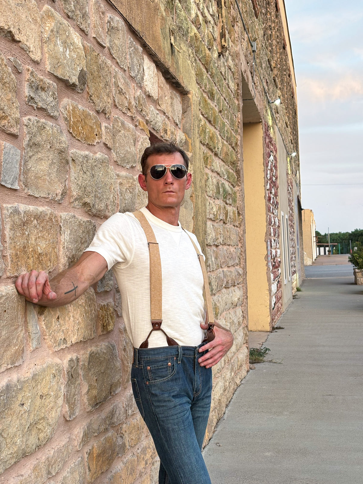 A man wearing a cream ribbed t-shirt, brown leather suspenders, and blue jeans leans against a rustic stone wall outdoors.