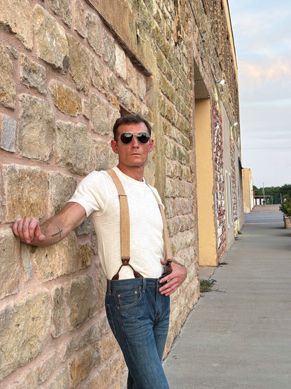 A man wearing a cream ribbed t-shirt, brown leather suspenders, and blue jeans leans against a rustic stone wall outdoors.