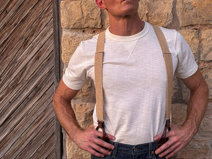 A man wearing beige suspenders with dark brown leather adjusters, styled over a white t-shirt and blue jeans, standing against a rustic stone wall.