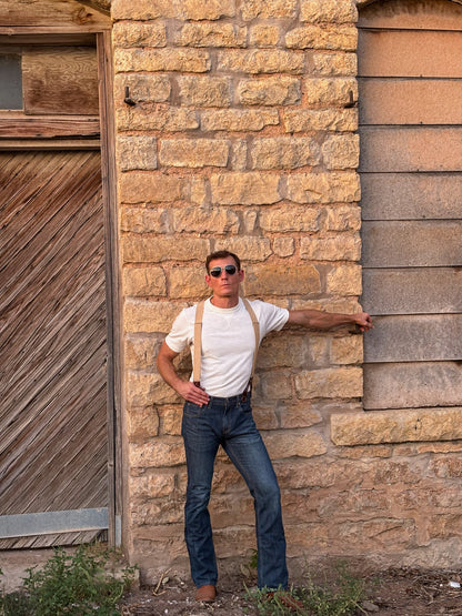 A man wearing a white t-shirt, blue jeans, and tan suspenders leans against a rustic stone wall next to a wooden door.