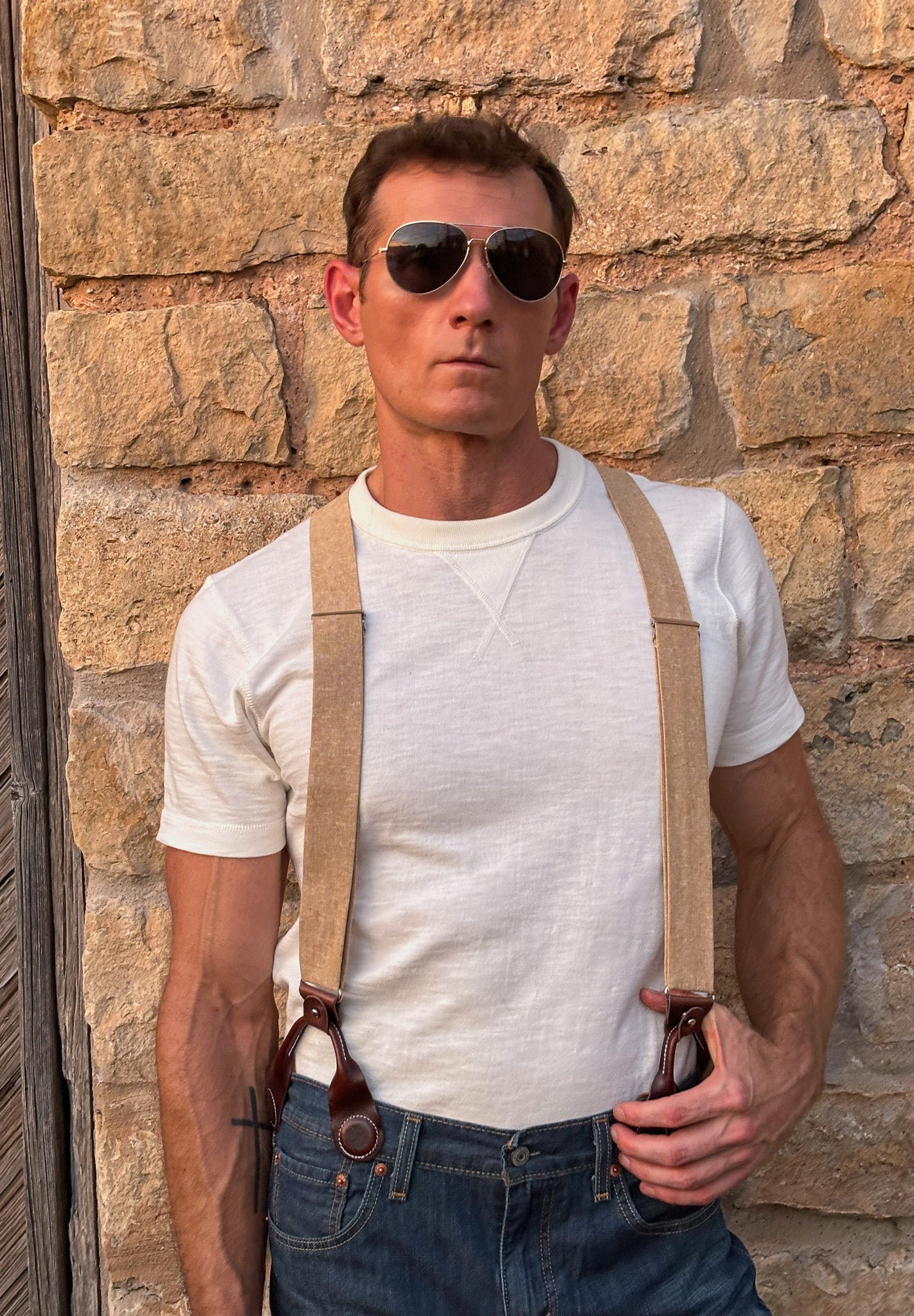 A man wearing a white crewneck t-shirt, beige  suspenders with brown leather hardware, and blue jeans, standing against a rustic stone wall.