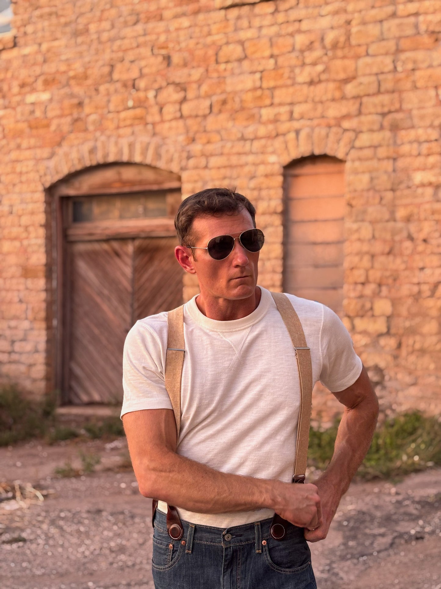 A man wearing aviator sunglasses, a white t-shirt, and tan suspenders with blue jeans, standing in front of a rustic brick building.