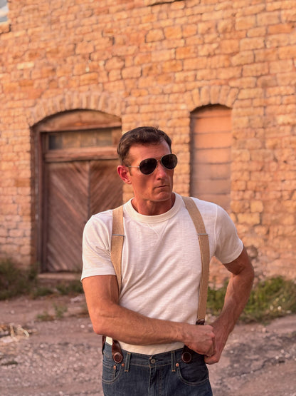 A man wearing aviator sunglasses, a white t-shirt, and tan suspenders with blue jeans, standing in front of a rustic brick building.