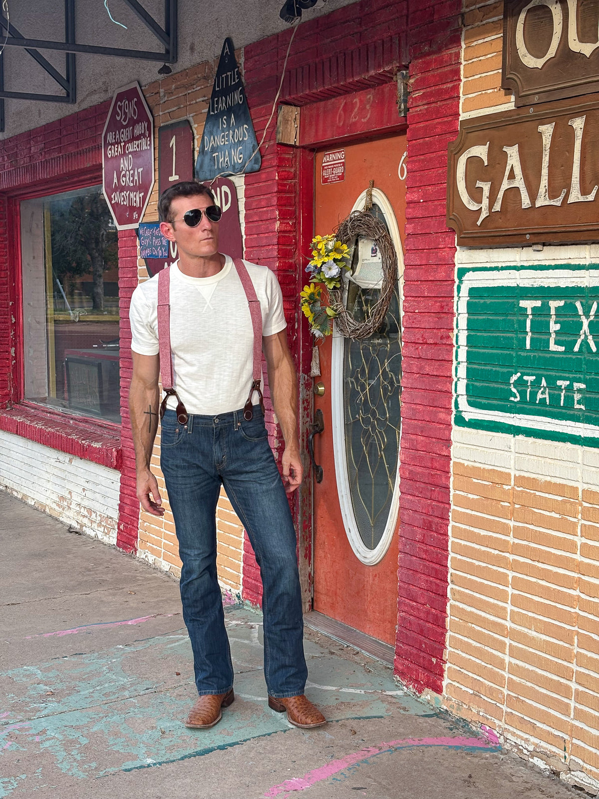 A man wearing a white t-shirt, dark blue jeans, red suspenders, and brown boots stands on a sidewalk in front of a brick storefront with a red door and vintage-style signs.