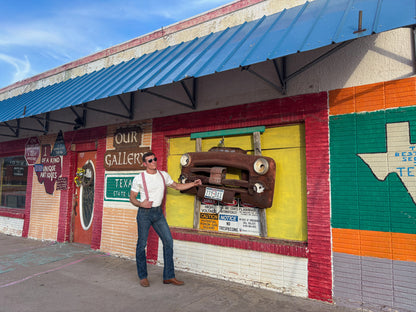 A man wearing sunglasses, a white shirt, and a red suspenders stands in front of a colorful building wall featuring a vintage car bumper decoration and a large painted Texas state sign.