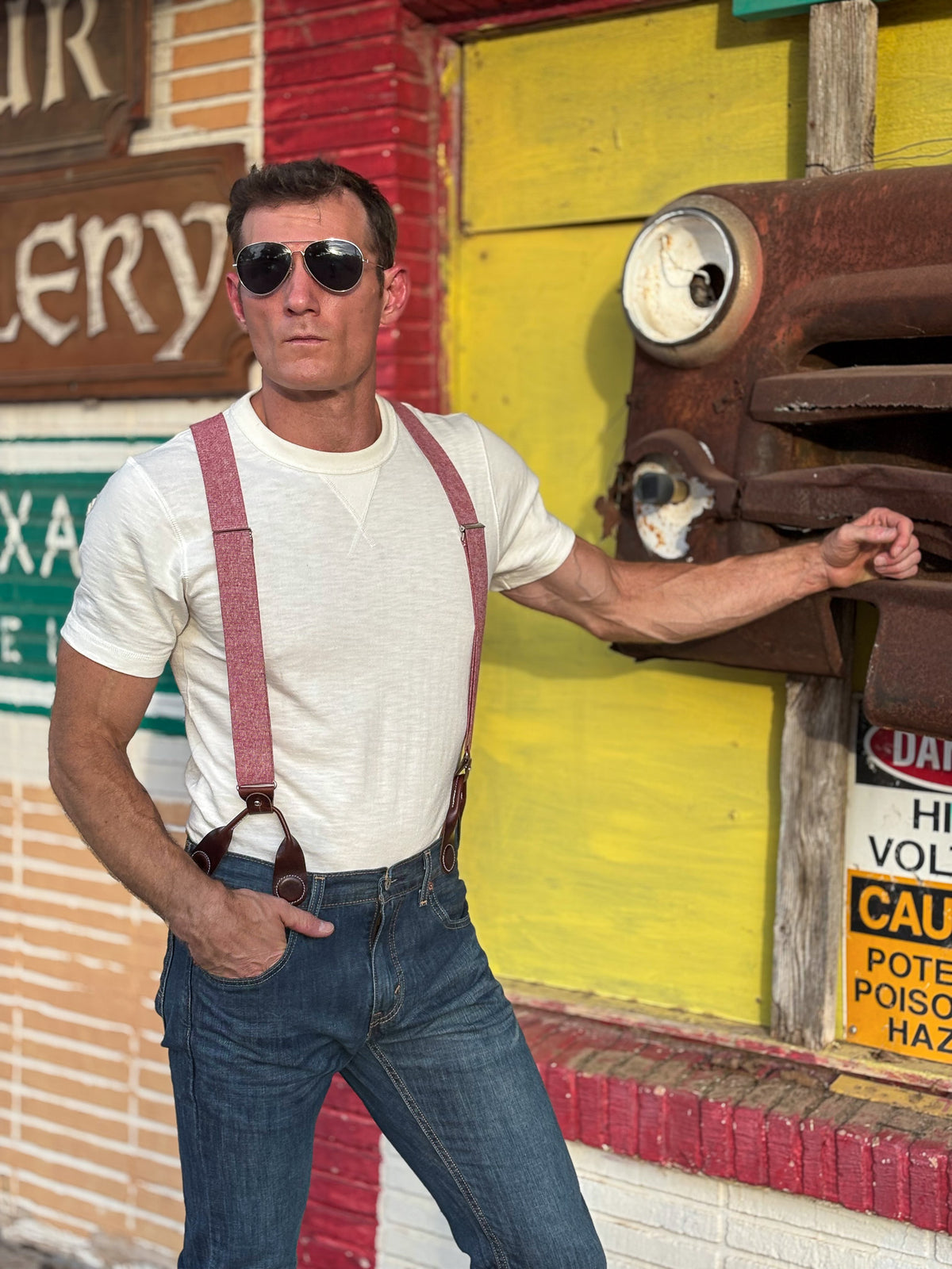 A man wearing a white t-shirt, red suspenders, and blue jeans stands in front of a rustic yellow and red building wall, touching a rusted metal fixture.