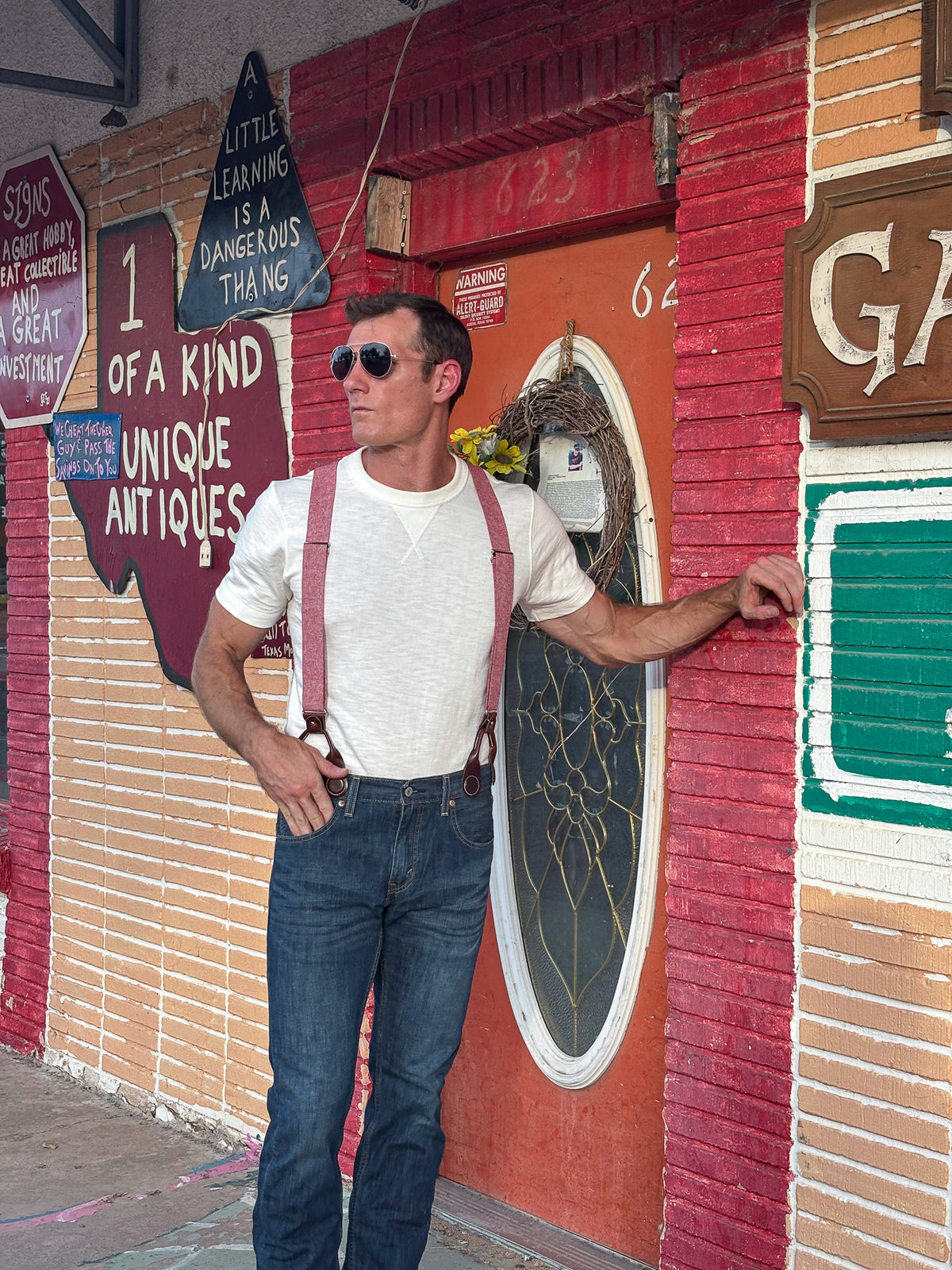 A man wearing a white t-shirt, blue jeans, and red suspenders stands in front of a colorful brick storefront with vintage signs.