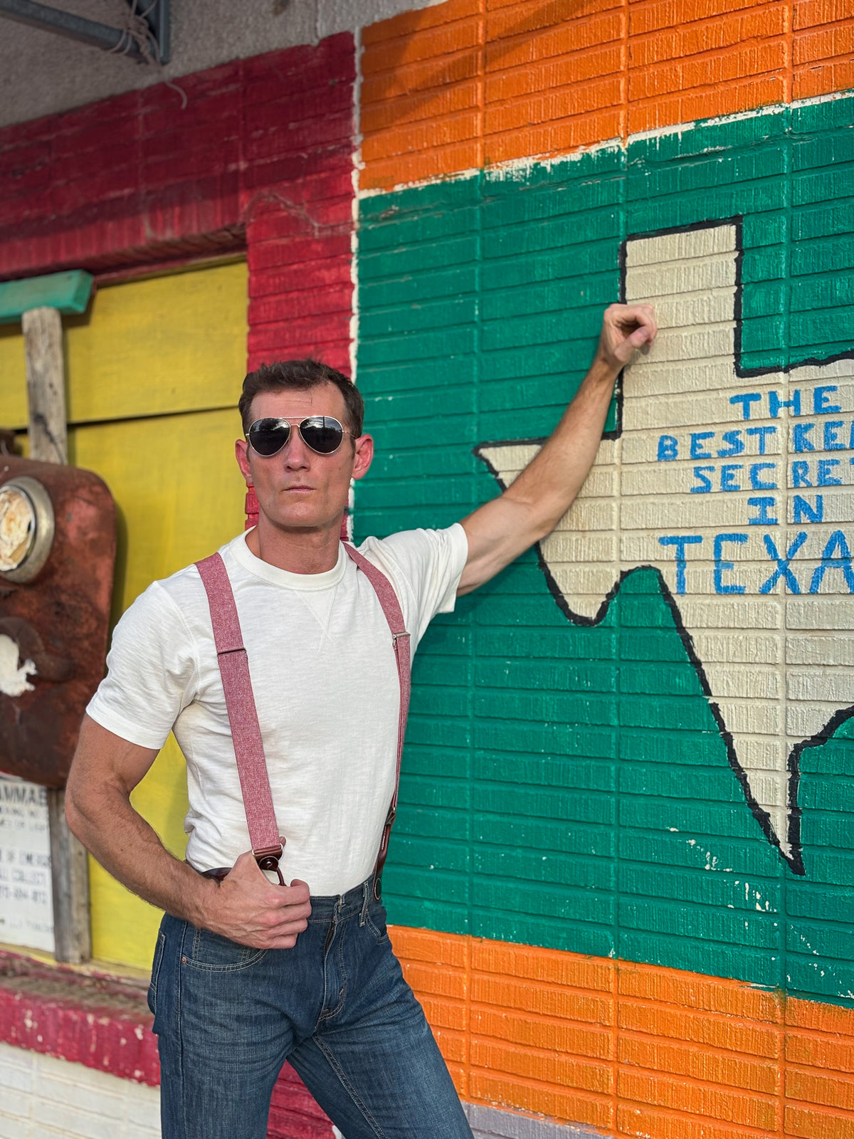 A man wearing sunglasses, a white t-shirt, and red suspenders poses in front of a colorful painted brick wall with a large Texas outline and blue text.