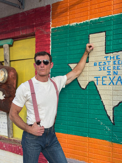 A man wearing sunglasses, a white t-shirt, and red suspenders poses in front of a colorful painted brick wall with a large Texas outline and blue text.