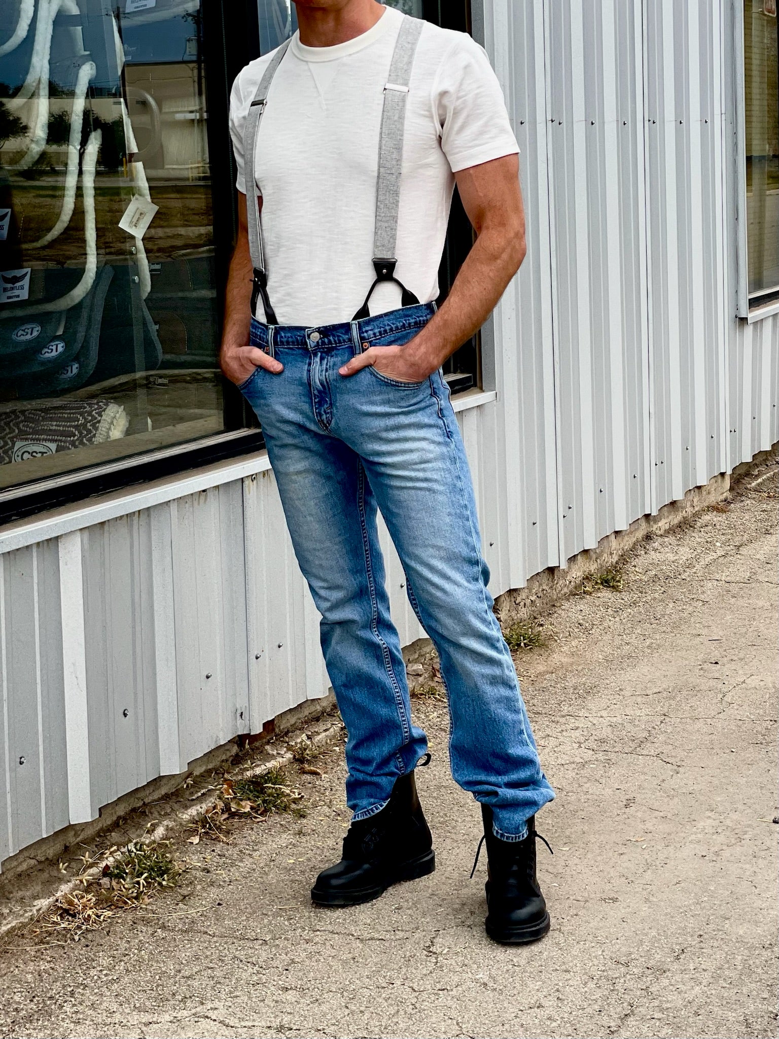 Man wearing steel gray button-on suspenders with a white t-shirt, blue jeans, and black work boots, standing against a corrugated metal building. A rugged yet refined accessory, perfect for weddings, vintage menswear, and workwear. Handmade in the USA.