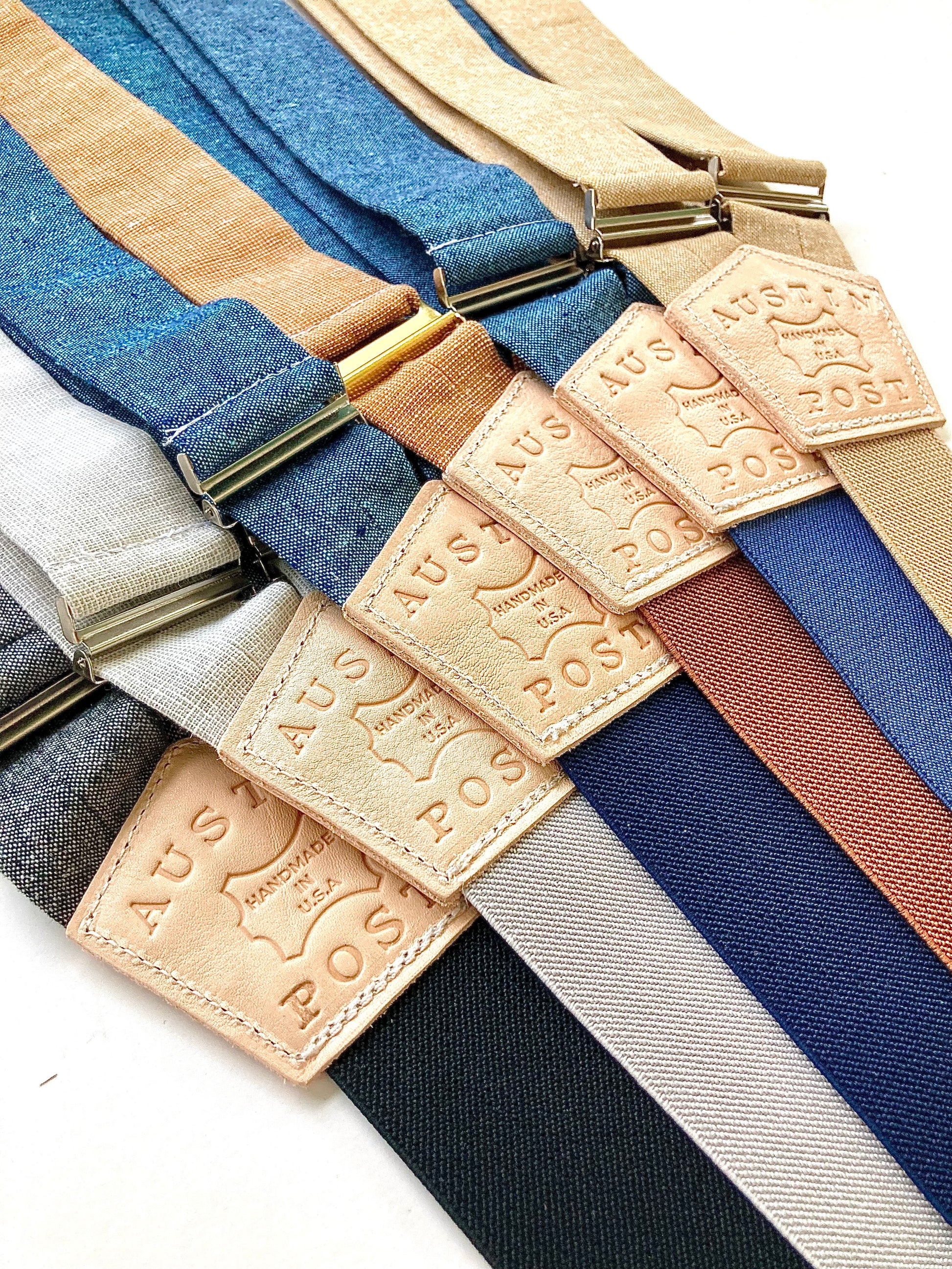 Assorted colors of Austin Post brand men’s suspenders with leather patches and brass hardware, handmade in the USA, displayed flat in a modern rustic style.