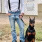 Man wearing black button-on suspenders with silver hardware over a white waffle-knit shirt and blue jeans, standing outdoors beside a Doberman pinscher; ideal for groomsmen, wedding parties, or vintage-inspired menswear looks.