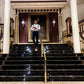 Man wearing blue linen magnetic suspenders at a formal wedding in Austin, walking down a grand staircase toward ballroom.