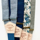 Close-up of five suspender strap colors in chambray blue, navy, midnight blue, teal floral, and sage green with Stratton Suspender Co. leather patch labels and mixed gold and silver adjusters.