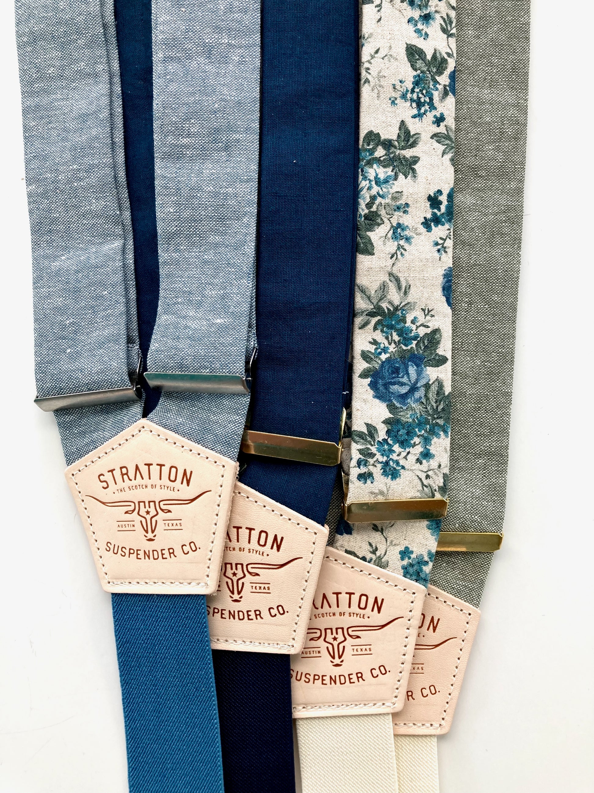 Close-up of five suspender strap colors in chambray blue, navy, midnight blue, teal floral, and sage green with Stratton Suspender Co. leather patch labels and mixed gold and silver adjusters.