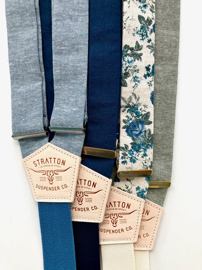 Close-up of five suspender strap colors in chambray blue, navy, midnight blue, teal floral, and sage green with Stratton Suspender Co. leather patch labels and mixed gold and silver adjusters.