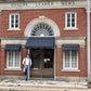 Man wearing handmade chocolate linen button-on suspenders and jeans, leaning casually outside historic Uvalde Leader-News building in Texas—vintage-inspired men's formalwear with Southern heritage style.