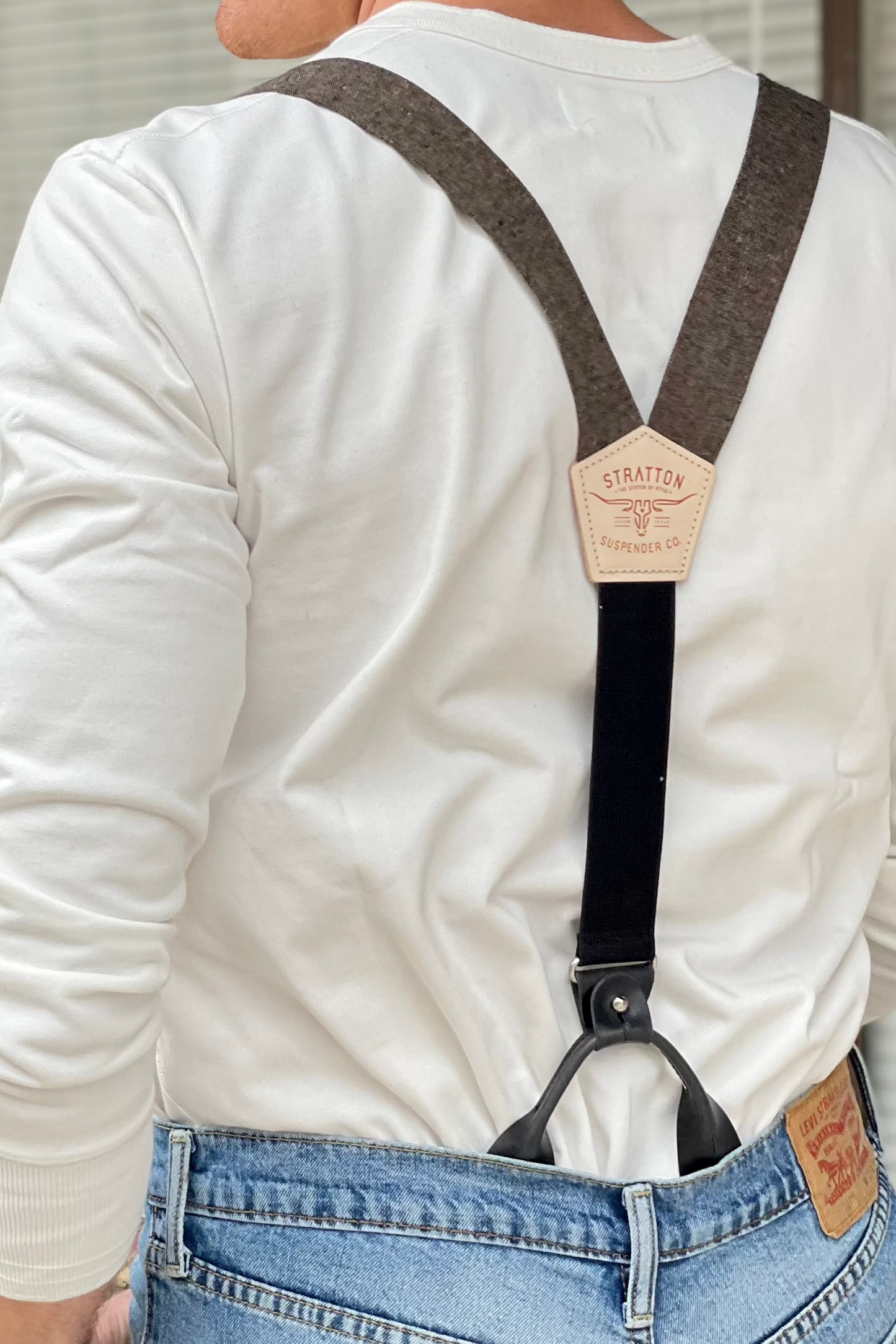 Back view of men's brown linen button-on suspenders with black leather attachments, featuring a Stratton Suspender Co. logo patch. Styled with a white Henley shirt and denim for a timeless, vintage-inspired look. Ideal for grooms, groomsmen, and classic wedding attire.