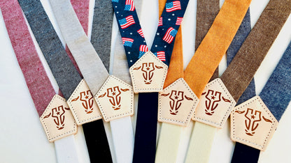 Children’s suspender collection in red, gray, beige, blue, brown, orange, and patriotic flag print, displayed with stitched leather back patches.