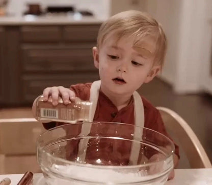 Young child wearing cream-colored suspenders while baking, pouring spices into a mixing bowl in a cozy home kitchen.