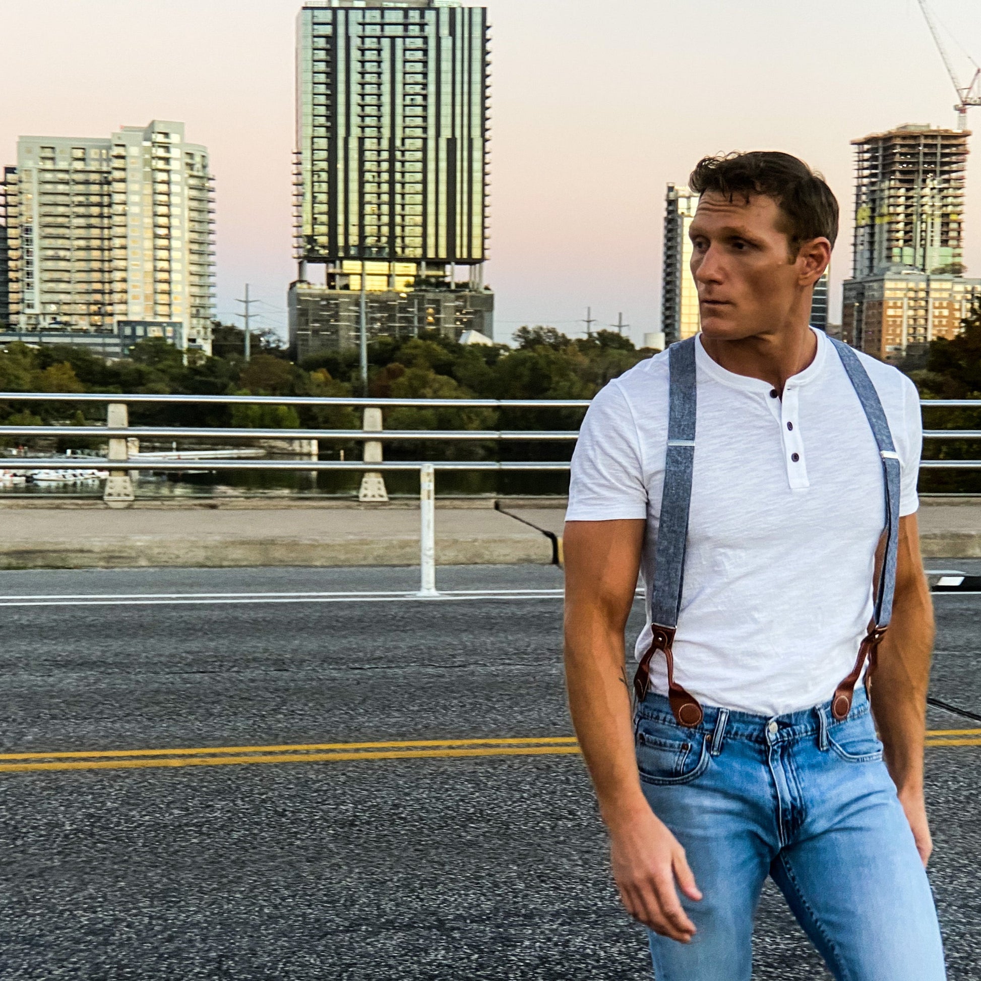 Man wearing Stratton Suspender Co. denim blue linen suspenders with cognac leather magnetic clasps, styled with a white henley and jeans against a modern cityscape. Handmade in the USA, these formal suspenders offer a polished alternative to clip-ons, perfect for weddings and menswear.