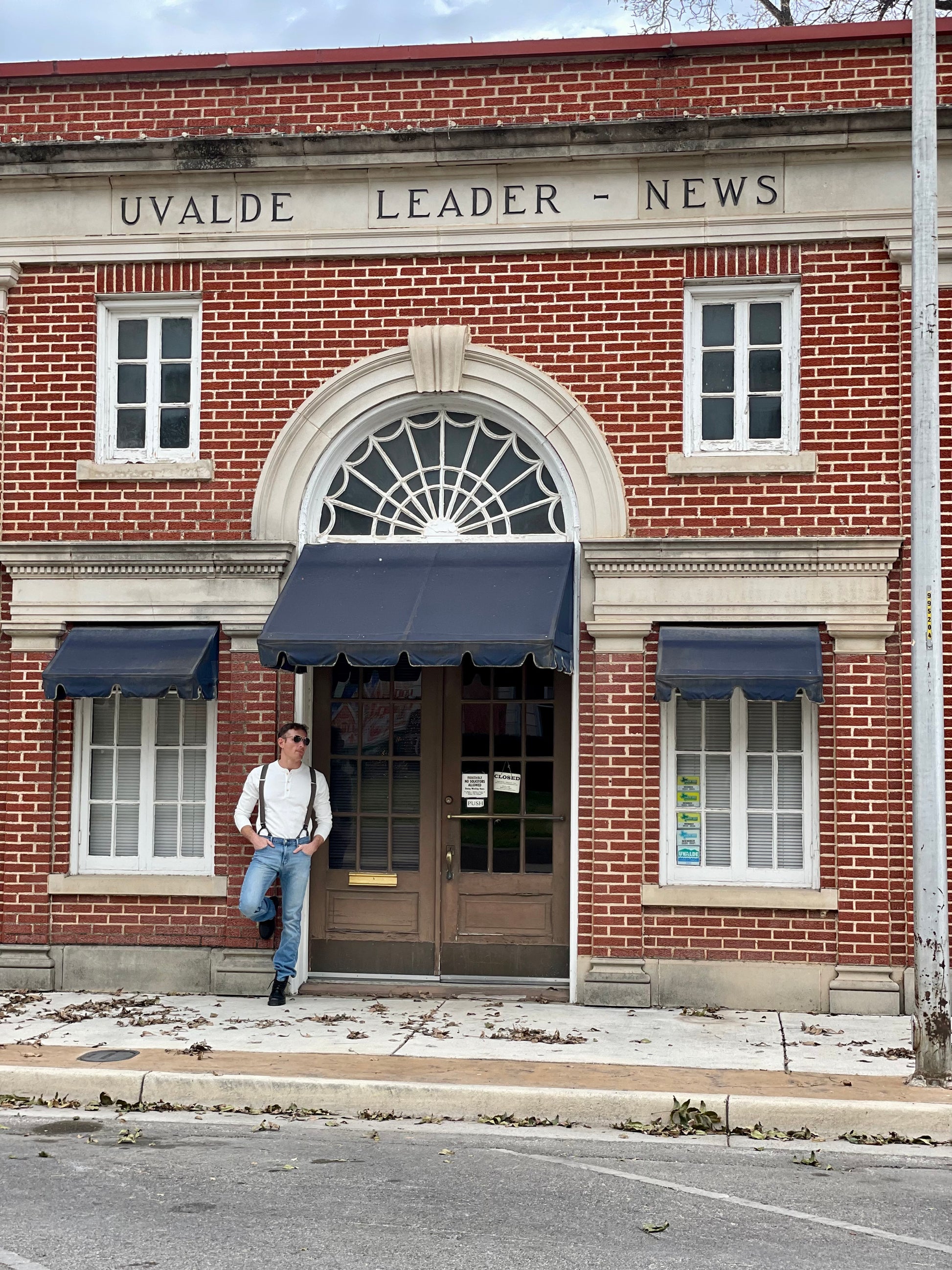 Man in espresso brown button-on suspenders leaning against historic red brick storefront in Texas, styled with jeans and a white Henley — rustic menswear and wedding accessory by Stratton Suspender Co.