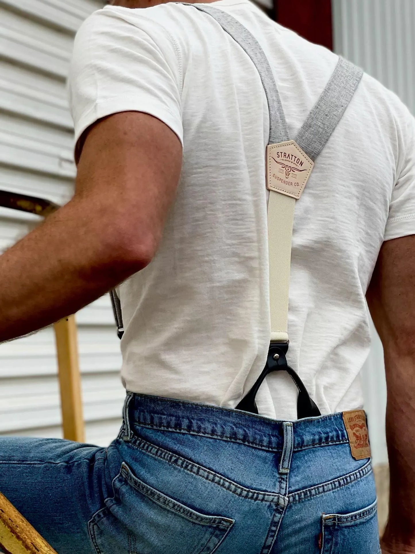 A close-up of a man wearing gray linen button-on suspenders with a natural leather patch, paired with a white t-shirt and classic denim jeans. The rugged yet refined style makes these suspenders perfect for groomsmen, rustic weddings, and vintage-inspired menswear.