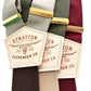 Close-up of Stratton brand suspenders in burgundy, olive green, and mushroom taupe with gold hardware and leather logo patch.