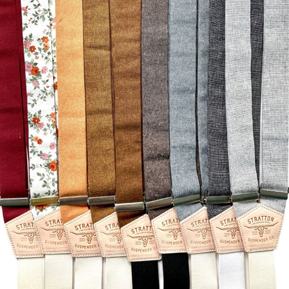 Assorted linen button-on suspenders in a range of solid, floral, and earth-tone fabrics with leather back patches, handmade in Texas for grooms, groomsmen, and heritage formalwear.