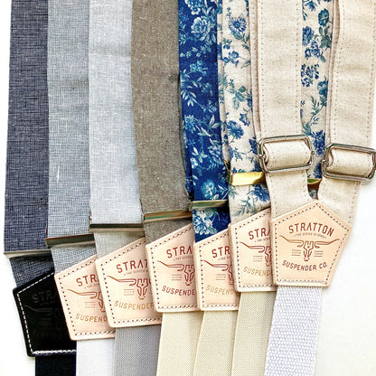 Collection of neutral linen suspenders from Stratton Suspender Co., featuring black, charcoal gray, olive, natural canvas, and vintage blue floral patterns. Handmade button-on braces with natural leather patchwork and polished metal hardware. Rustic men’s wedding and heritage menswear accessories.