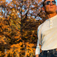 Man wearing ivory beige linen button-on suspenders with brown leather tabs and henley shirt, looking out in front of fall foliage. Rustic Texas menswear perfect for weddings or vintage-inspired formal looks.