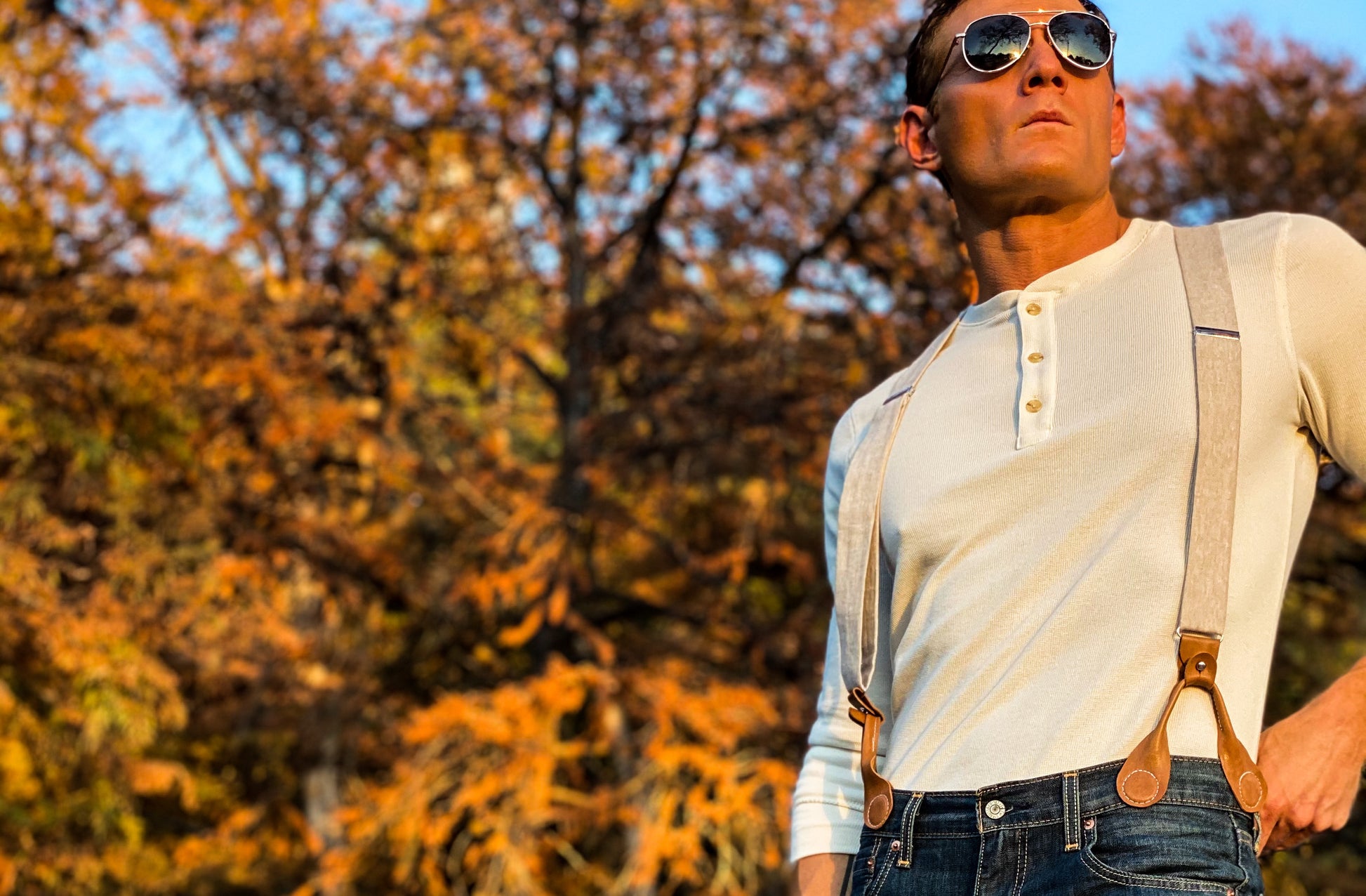 Man wearing ivory beige linen button-on suspenders with brown leather tabs and henley shirt, looking out in front of fall foliage. Rustic Texas menswear perfect for weddings or vintage-inspired formal looks.