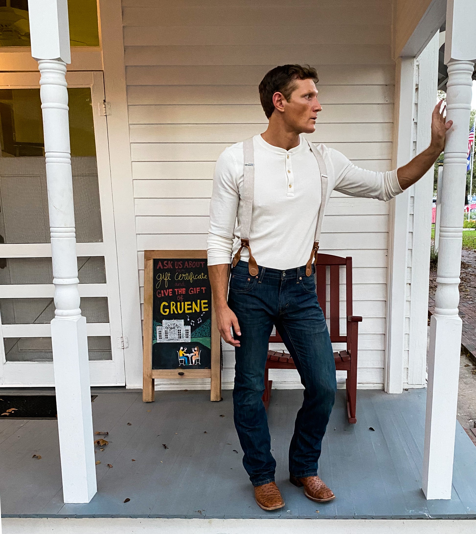 Man wearing handmade ivory beige linen suspenders with brown leather button tabs, styled with dark jeans and a henley shirt, standing on the porch of Gruene Hall in New Braunfels, Texas. Southern rustic wedding or heritage menswear look.