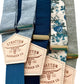 Flat lay of linen suspenders in sky blue, navy, deep blue, floral teal, and sage green with gold and silver adjusters—handmade by Stratton Suspender Co. for weddings, groomsmen, and vintage menswear styling.