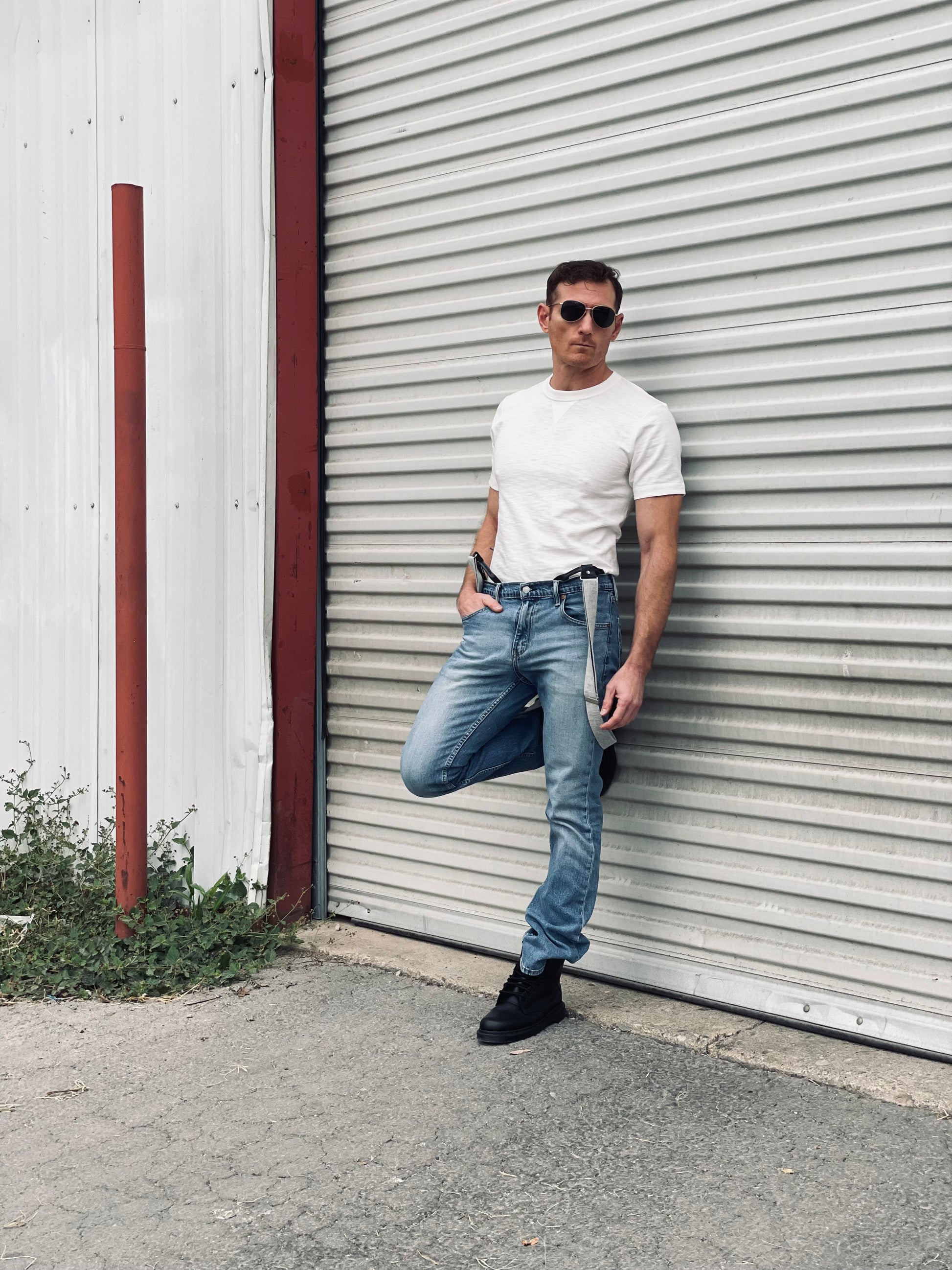 Man wearing light gray canvas suspenders with leather loops, blue jeans, and a white tee, standing outdoors against a corrugated metal wall — rugged men’s casual suspenders for everyday or workwear style.