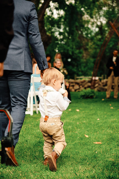 Young ring bearer wearing children’s suspenders with leather accents at an outdoor wedding, holding hands with an adult in a navy suit.