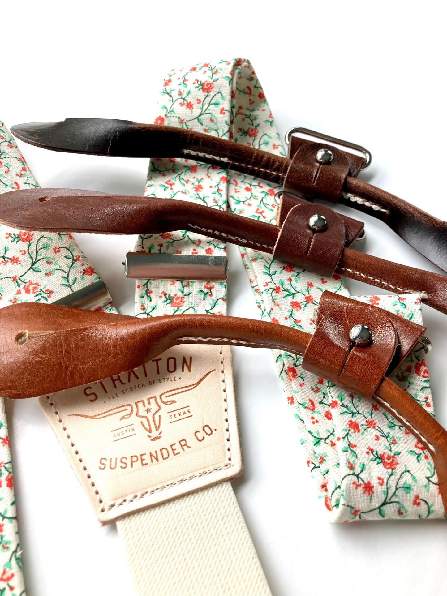 Close-up of rustic orange and green floral suspenders with brown leather button-on loops and cream elastic back, handmade by Stratton Suspender Co. for groomsmen and rustic wedding attire.