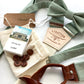 Sage green linen wedding suspenders with bow tie, leather button-on ends, sewing kit, and custom name tag—groomsmen gift set handmade for rustic weddings.