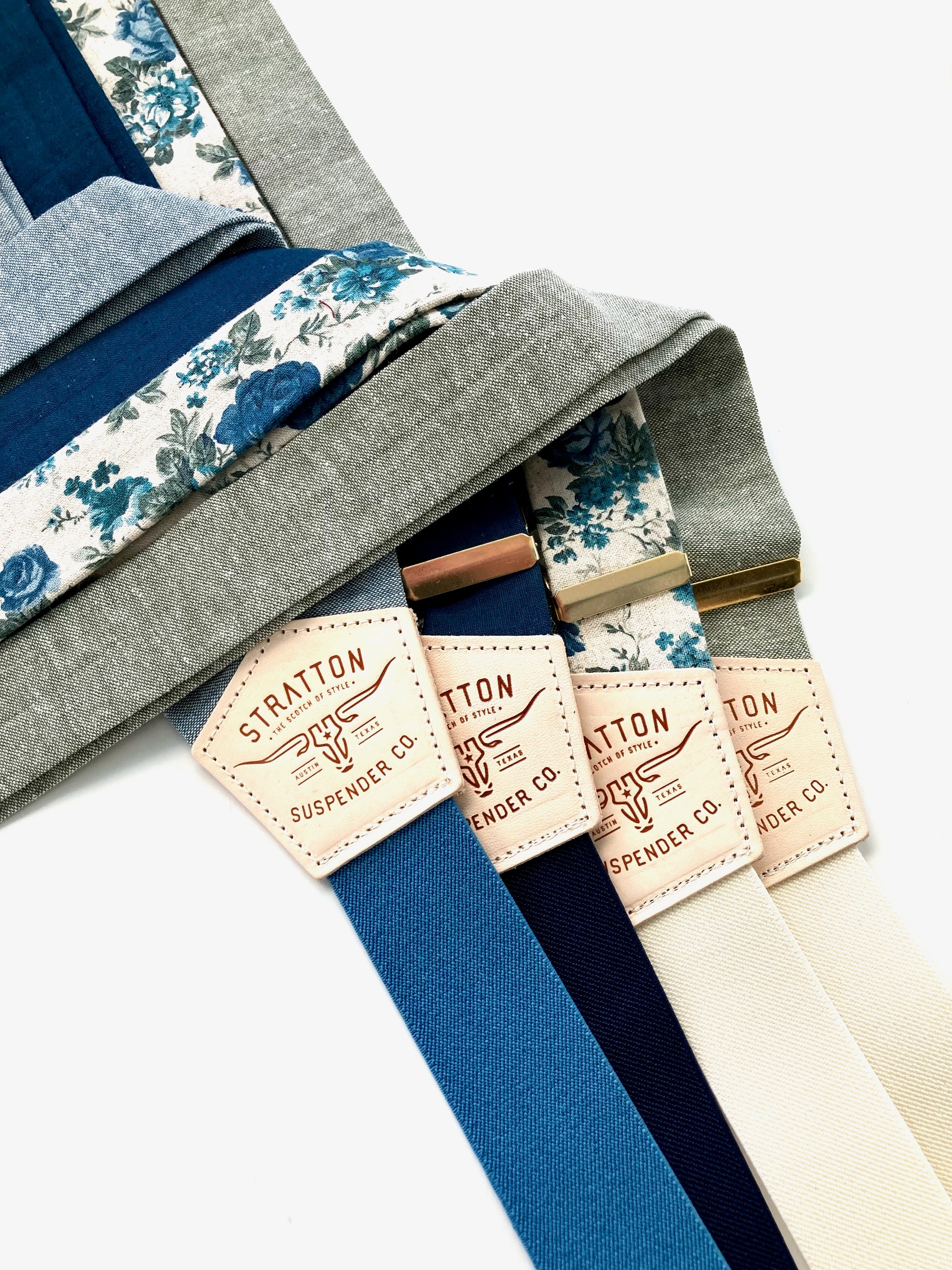 Men’s linen suspenders in a soft palette of sky blue, midnight blue, teal floral, and sage green with cream and ivory back straps and metal adjusters.