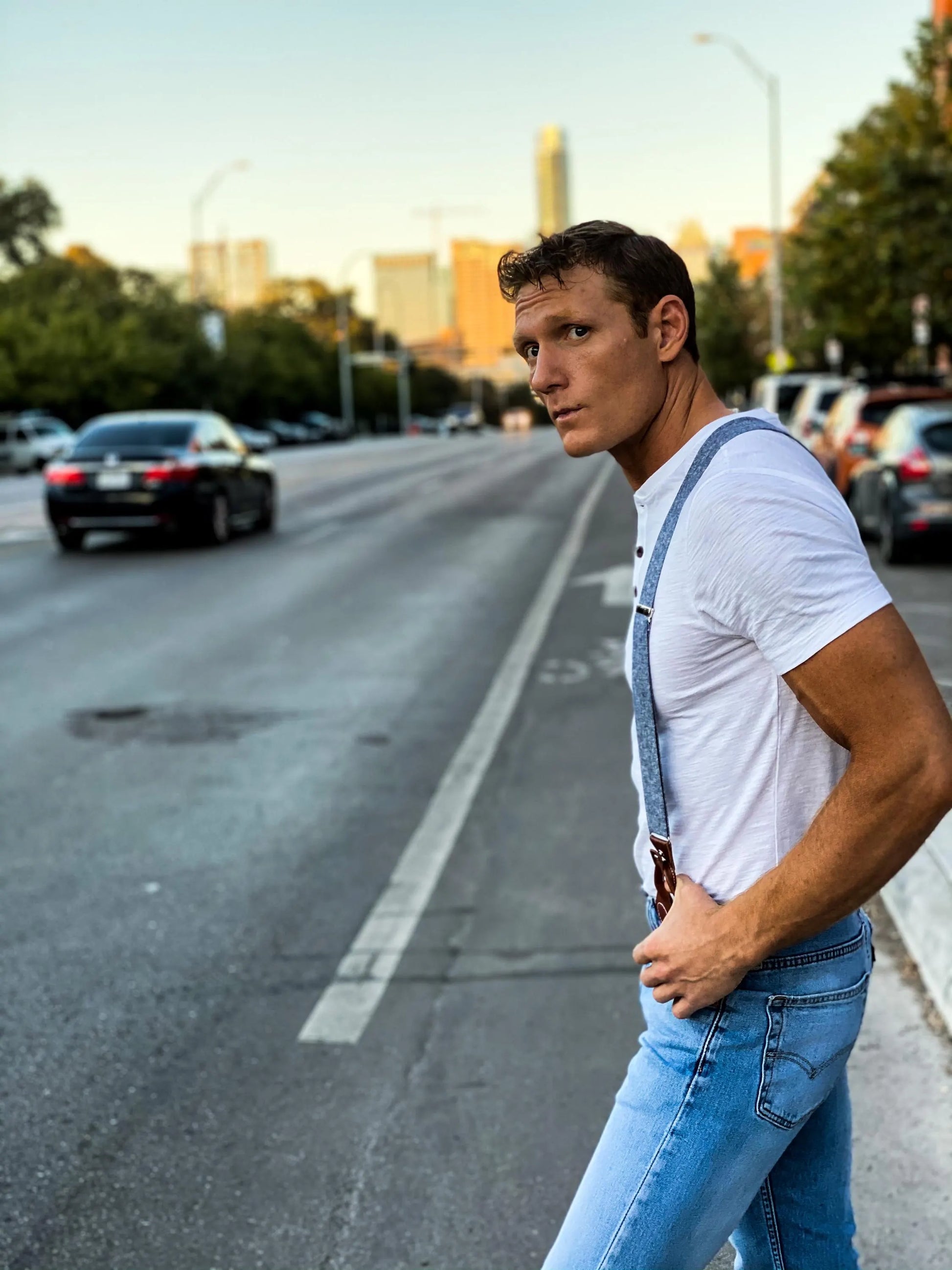 Model wearing Stratton Suspender Co. denim blue linen suspenders with button ends and a white Henley tee, walking on South Congress Street in Austin, Texas with the downtown skyline in the background.