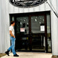 Man in vintage-inspired suspenders and jeans walks past a rustic Texas farm store, showcasing handmade wedding and groomsmen accessories from Stratton Suspender Co.