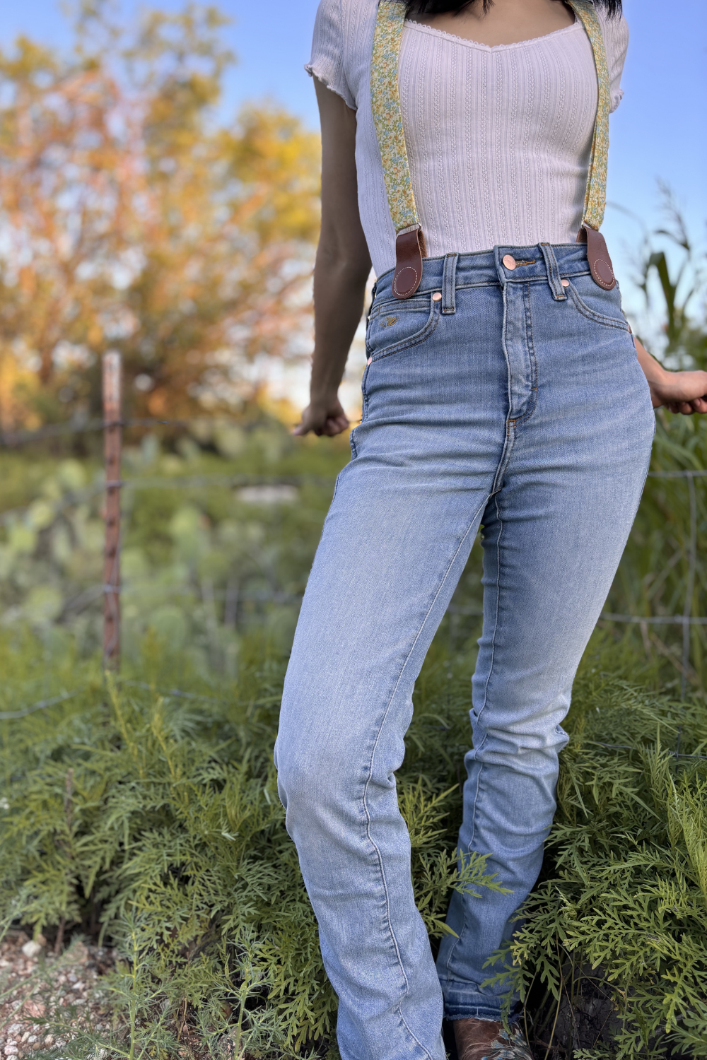Close-up of floral women’s magnetic clasp suspenders with leather tabs, styled with blue jeans and a white knit top.