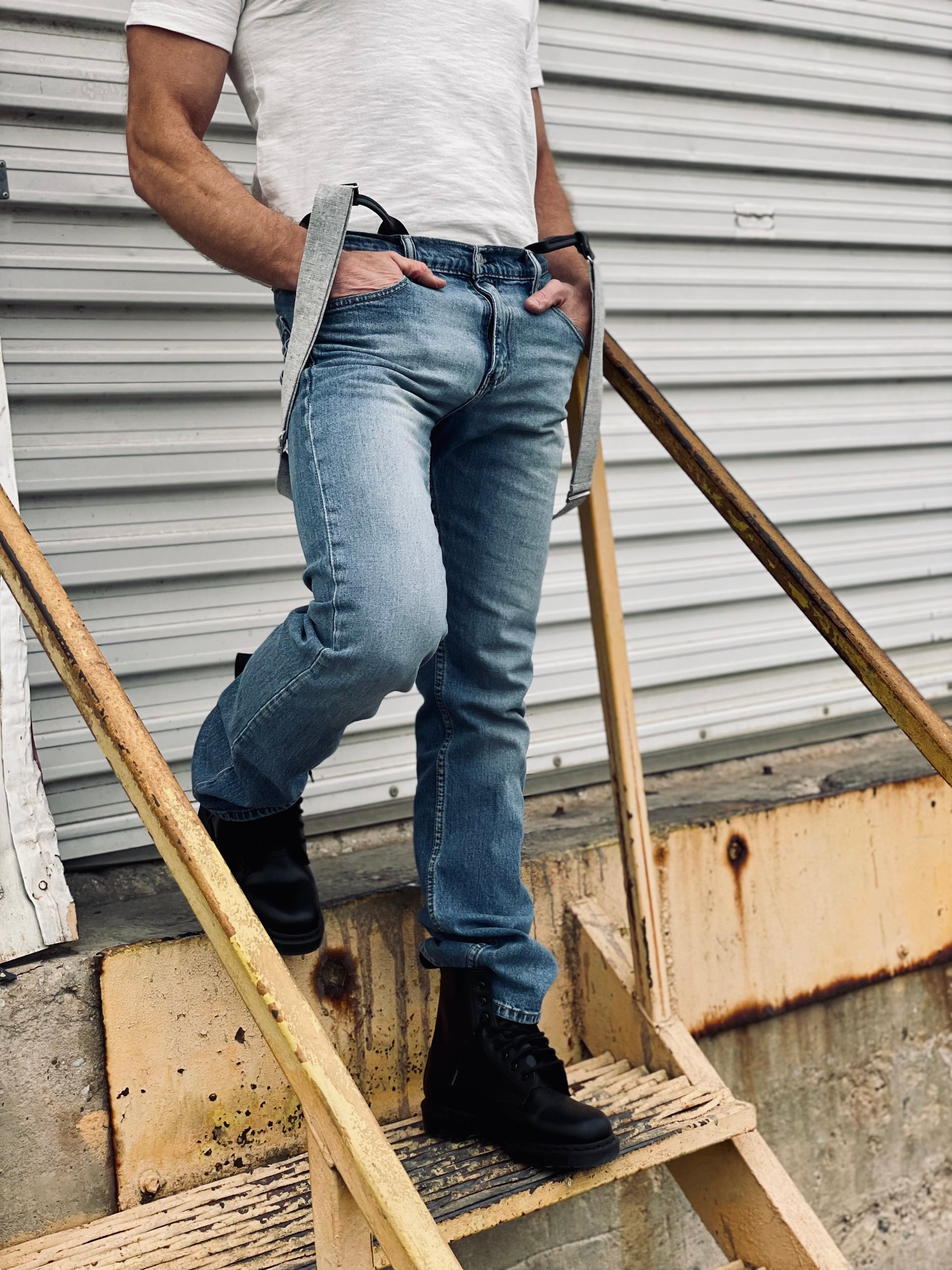 Man standing on industrial staircase wearing vintage charcoal linen button-on suspenders with black leather ends, paired with a white tee, light-wash Levi’s jeans, and black combat boots—handmade suspenders for weddings, groomsmen, and everyday classic menswear.
