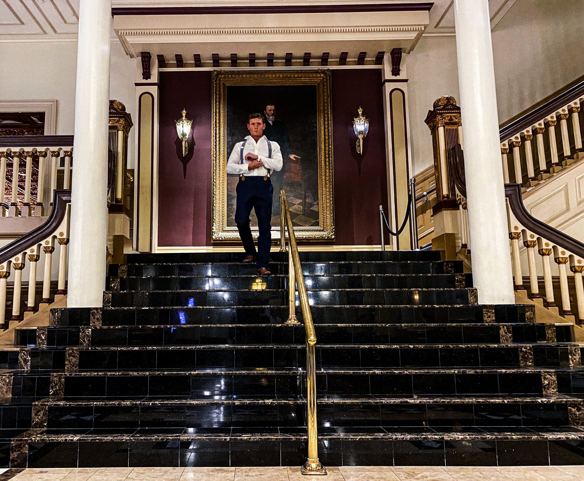 Model in formal navy trousers and suspenders stands on a grand staircase in a vintage hotel lobby, showcasing Stratton Suspender Co. wedding and event menswear.