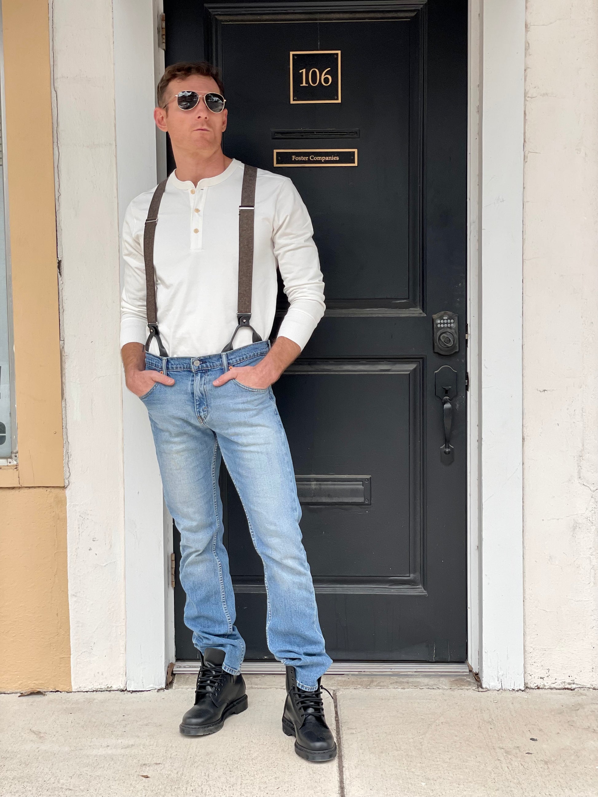 Man wearing a Vintage White Henley and brown wool suspenders by Stratton Suspender Co., standing confidently in front of a black door. Paired with denim and black boots, this rugged heritage menswear look embodies classic American workwear and craftsmanship.